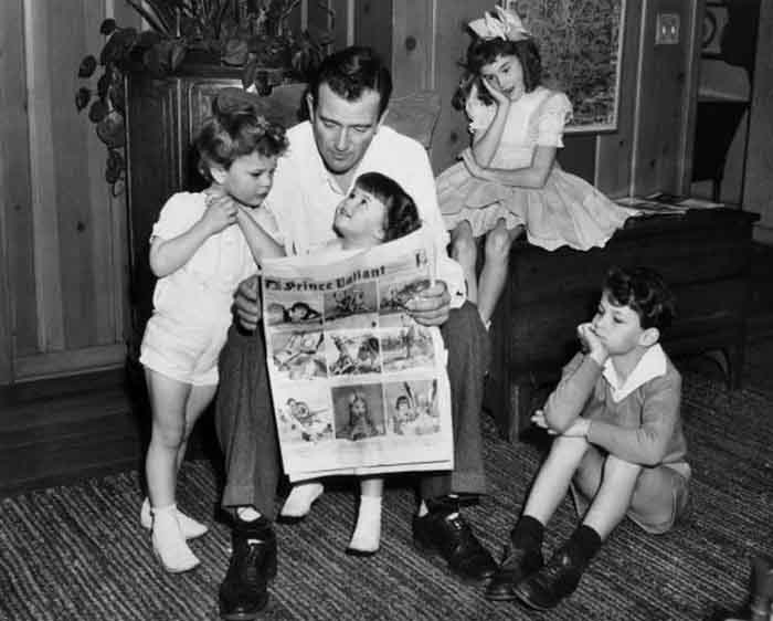 1942:  American actor John Wayne sitting on a chair and reading a Prince Valiant comic with his four children, Hollywood, California.  From left: Patrick, Melinda, Toni, and Michael.  Patrick and Melinda are leaning on their father, while the other two children listen from a desk and the floor.  (Photo by Hulton Archive/Getty Images)